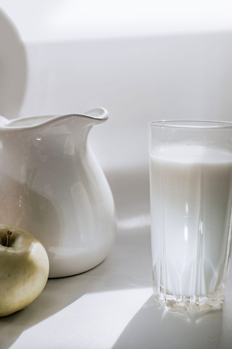 A serene still life image of milk in a glass next to a green apple and ceramic pitcher.